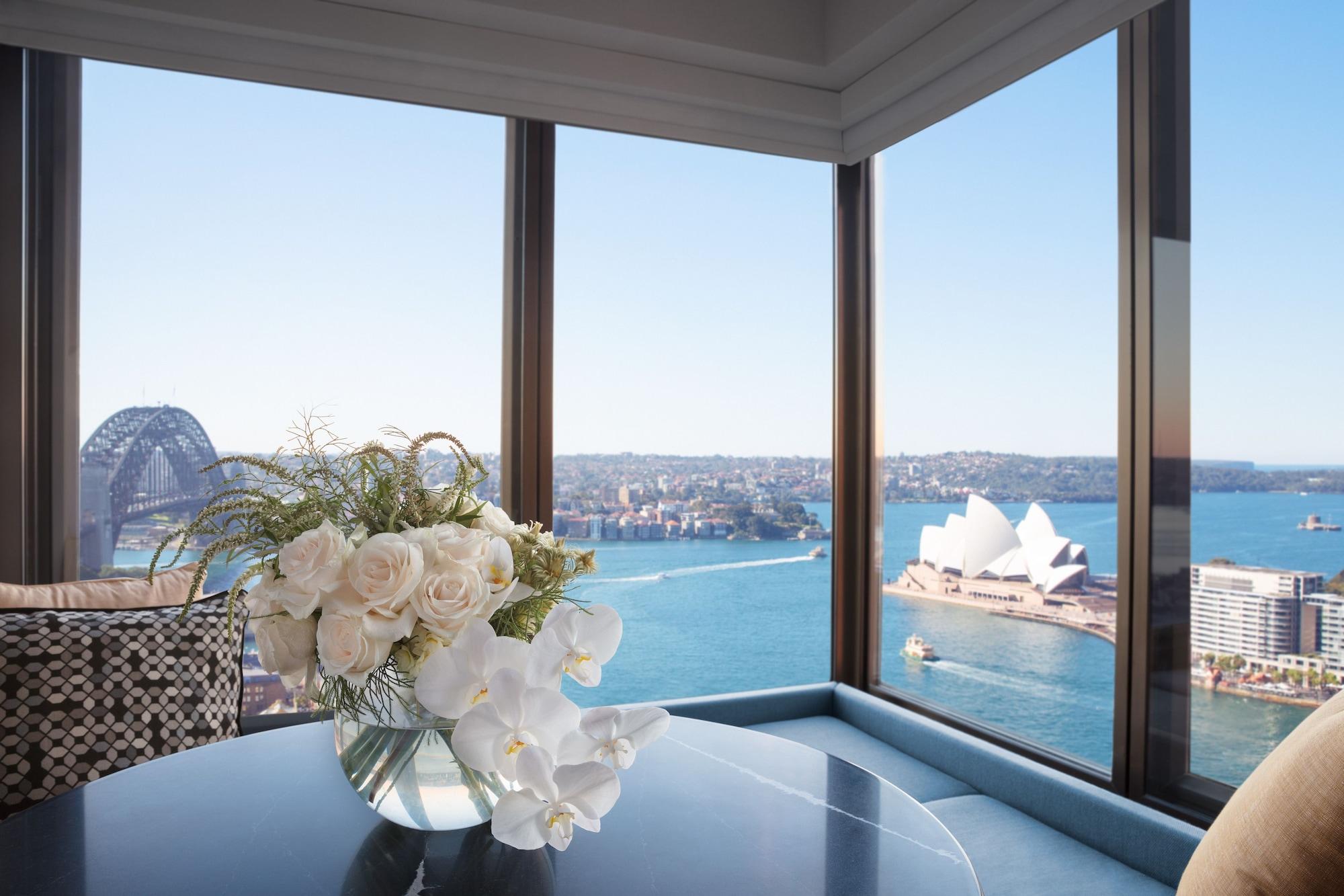 Four Seasons Hotel Сидней Экстерьер фото The photo shows an elegant interior space with large windows that provide a stunning view. In the foreground, there is a stylish table topped with a beautiful floral arrangement consisting of white roses and orchids in a clear vase. Through the windo