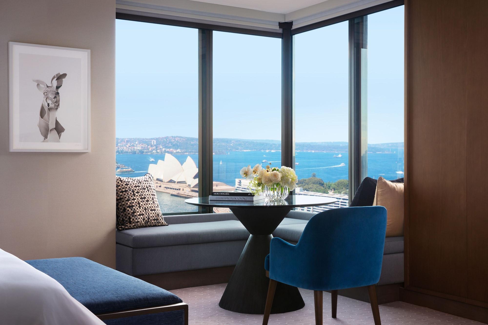 Four Seasons Hotel Сидней Экстерьер фото The photo shows a stylish hotel room with a large window offering a view of the water and the Sydney Opera House in the distance. There is a small, modern dining table with a vase of flowers on it, situated near the window. A comfortable blue chair i
