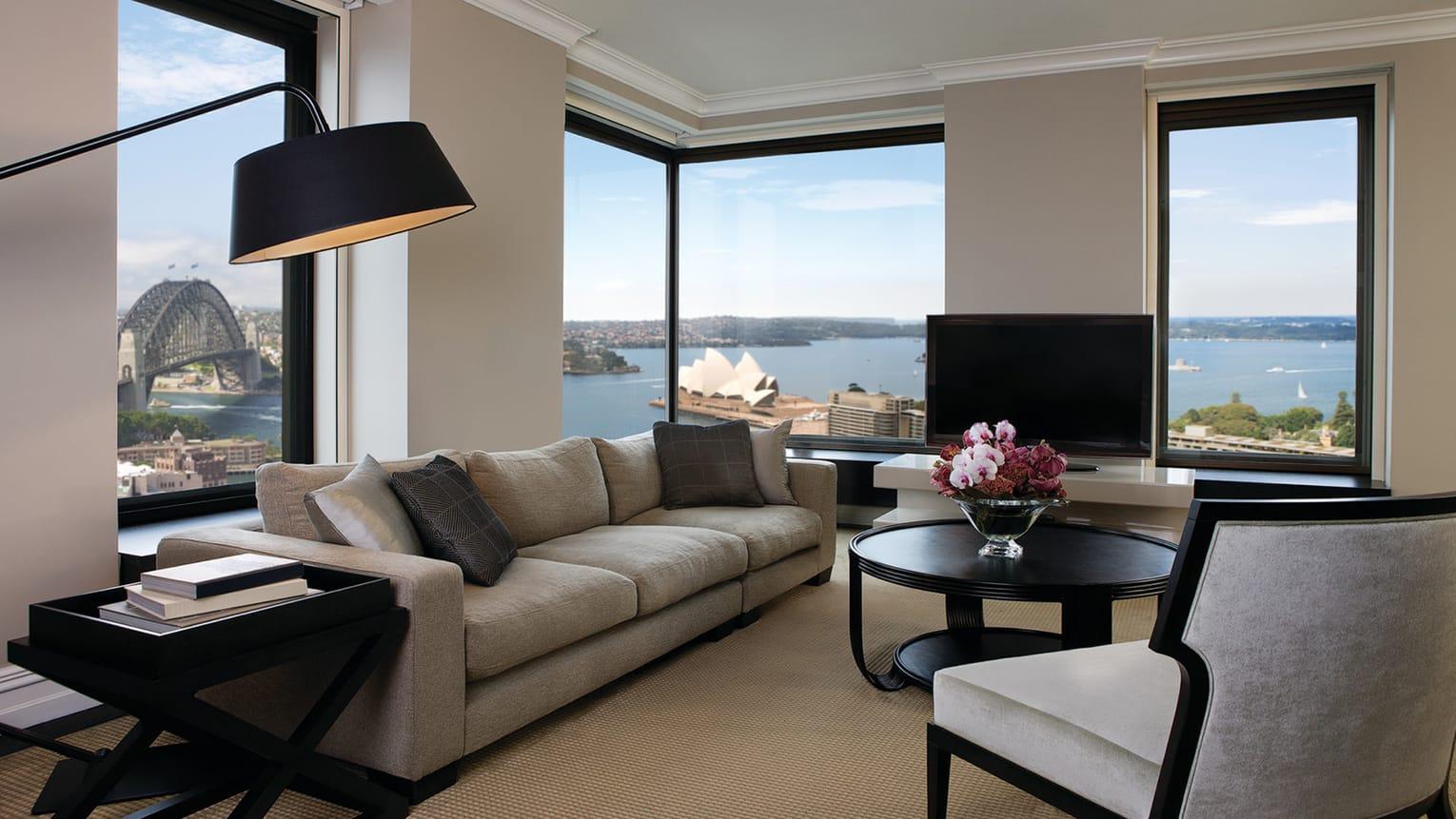 Four Seasons Hotel Сидней Экстерьер фото The photo shows a stylish and modern living room with large windows offering a view of a body of water and nearby buildings. The room features a comfortable, light-colored sofa and a round coffee table with a floral centerpiece. A flat-screen televis
