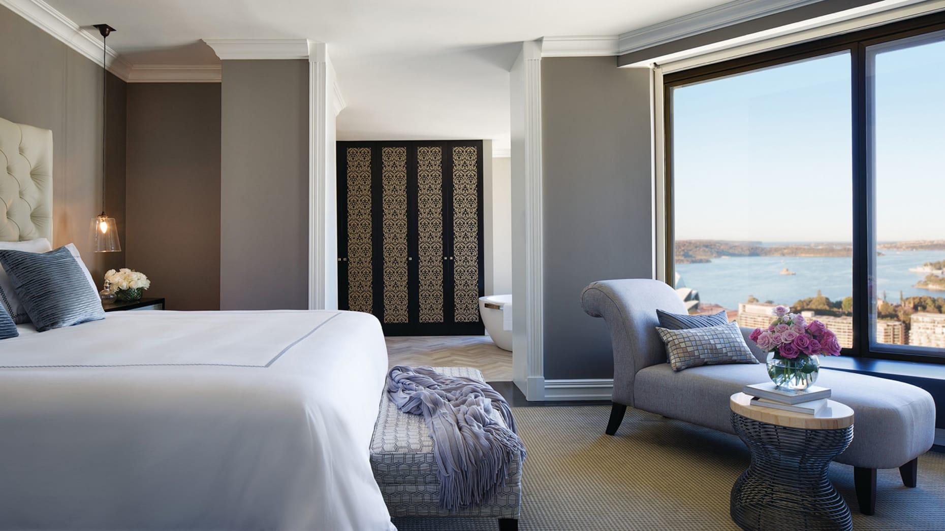 Four Seasons Hotel Сидней Экстерьер фото The photo shows a stylish hotel room featuring a large bed with crisp white linens. To the left, there is a comfortable seating area with a gray accent chair and a small round table topped with a vase of flowers. The room has a modern design, charact