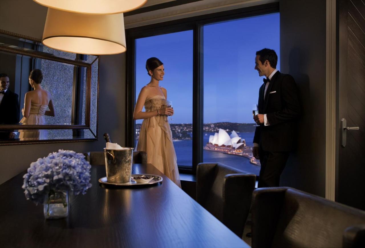 Four Seasons Hotel Сидней Экстерьер фото The photo depicts a stylish evening scene where a woman in an elegant dress and a man in a formal suit are engaged in conversation near a large window. They are in a modern interior space, with a table that has a bottle of champagne and a glass. The 