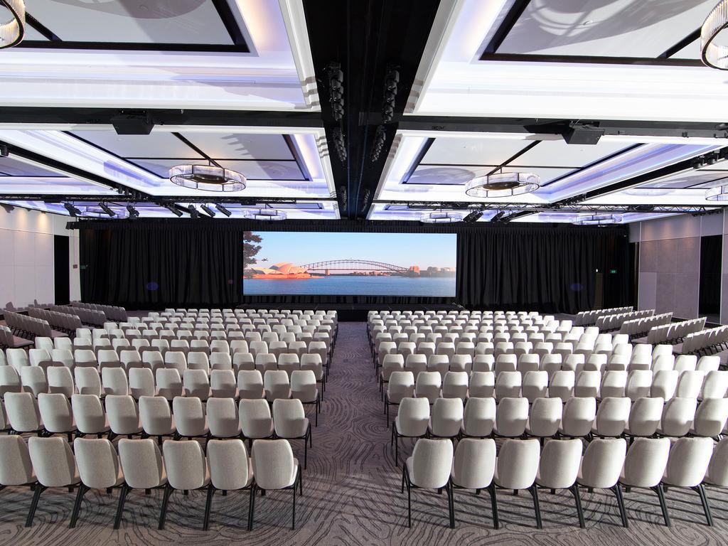 Four Seasons Hotel Сидней Экстерьер фото The photo depicts a large conference hall or auditorium filled with rows of white chairs arranged in a semi-circle facing a central stage or screen. The room has a modern design, featuring stylish ceiling lights and a sleek, contemporary aesthetic. A
