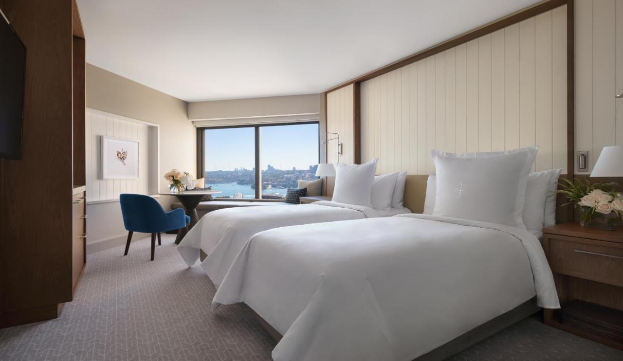 Four Seasons Hotel Сидней Экстерьер фото The photo shows a contemporary hotel room featuring two neatly made beds with white linens. There is a cozy blue chair positioned near a table, and a vase with flowers on the table adds a touch of decor. The large window offers a view of the outside,